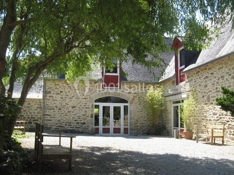 Location salle Douarnenez (Finistère) - La Ferme de Trézulien #3 Cour d'une maison en pierre avec des fenêtres rouges, entourée d'arbres et de bancs en bois.