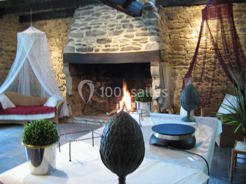 Location salle Douarnenez (Finistère) - La Ferme de Trézulien #18 Cheminée en pierre allumée dans une pièce rustique avec table décorée et fauteuil sous un voile.