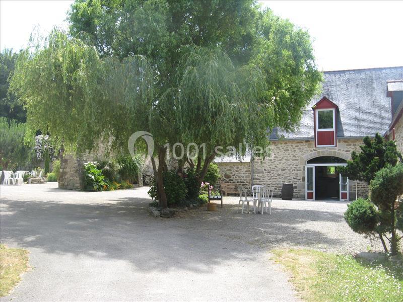 Location salle Douarnenez (Finistère) - La Ferme de Trézulien #4 Cour avec un grand arbre, des chaises blanches et une maison en pierre avec une porte et des fenêtres rouges.