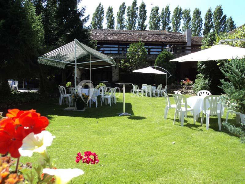Jardin avec pelouse, tables et chaises blanches sous des parasols, entouré de verdure et d'arbres.