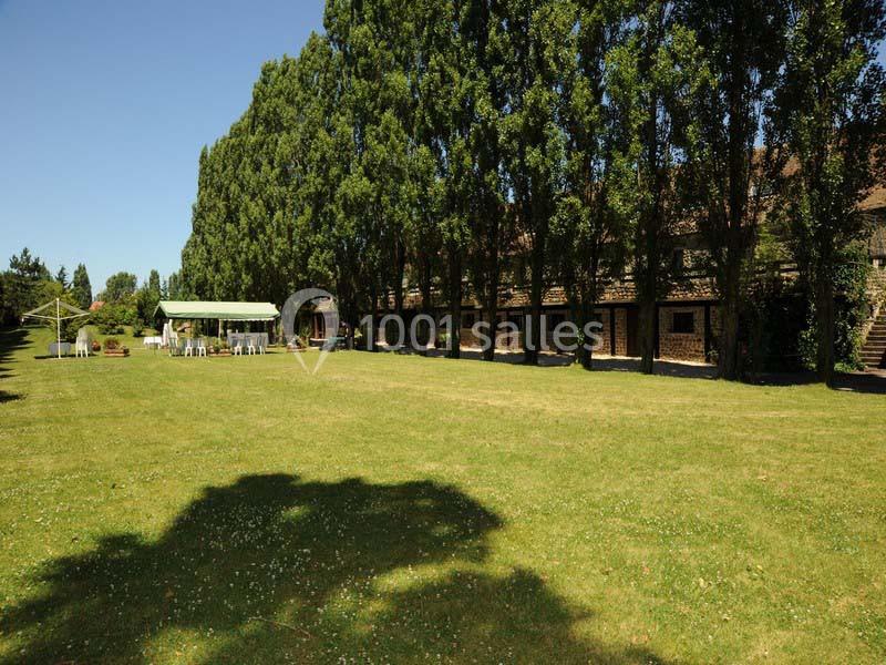 Grand jardin avec pelouse, arbres alignés et une structure couverte avec des tables et chaises blanches.