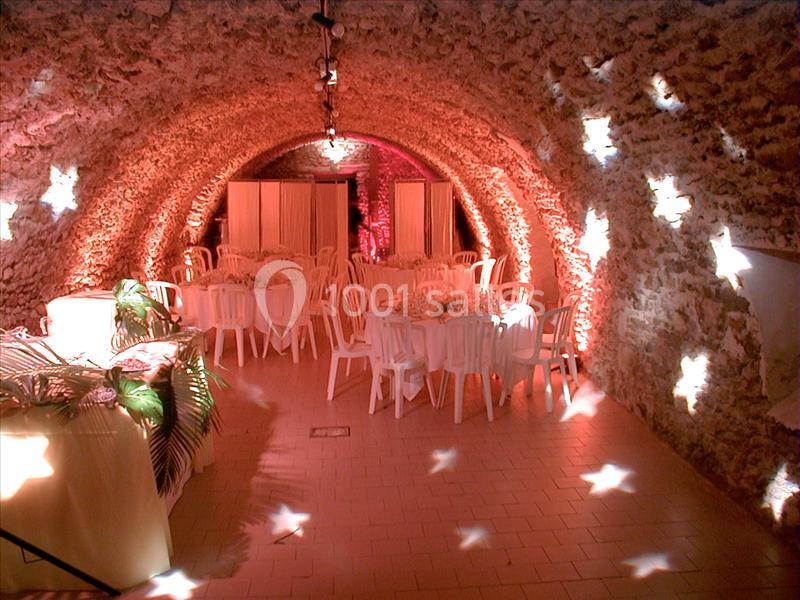 Salle voûtée en pierre éclairée par des lumières tamisées, aménagée avec des tables rondes et des chaises blanches.
