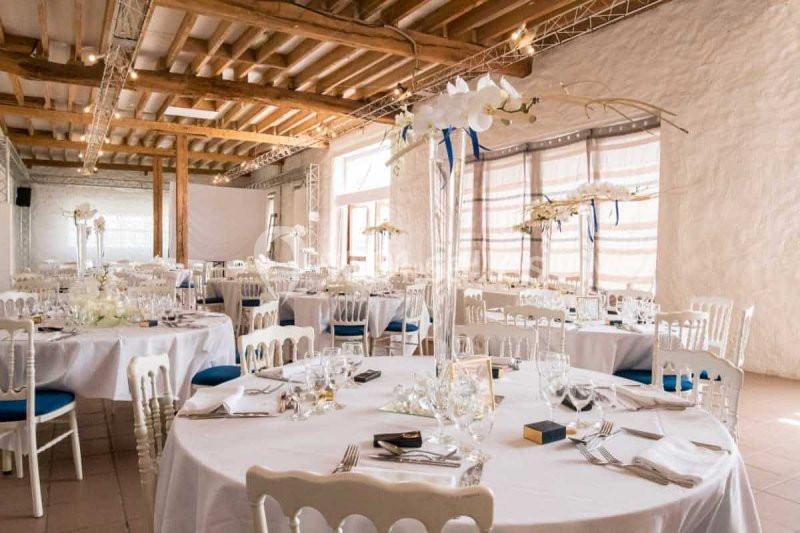 Salle de réception lumineuse avec tables rondes dressées, nappes blanches, chaises en bois et décorations florales.