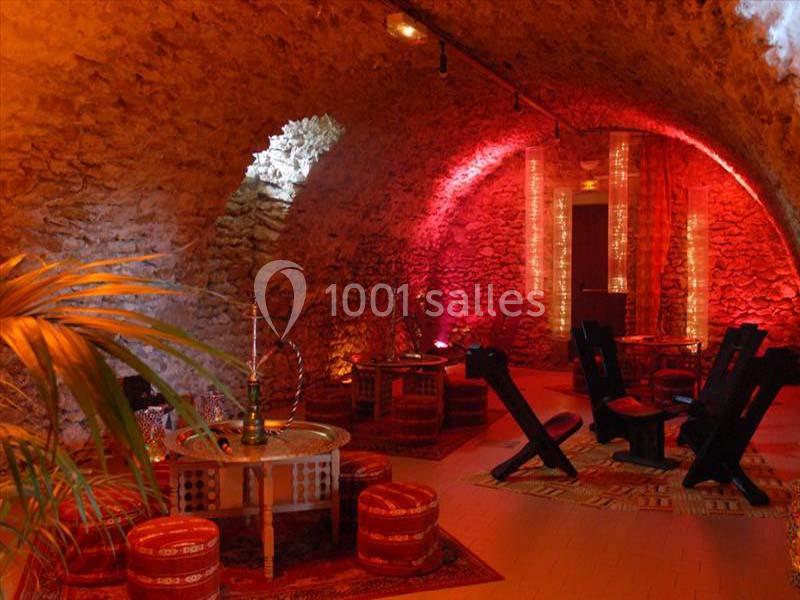 Salle voûtée en pierre avec éclairage rouge, tables basses, poufs et narguilés disposés dans une ambiance orientale.