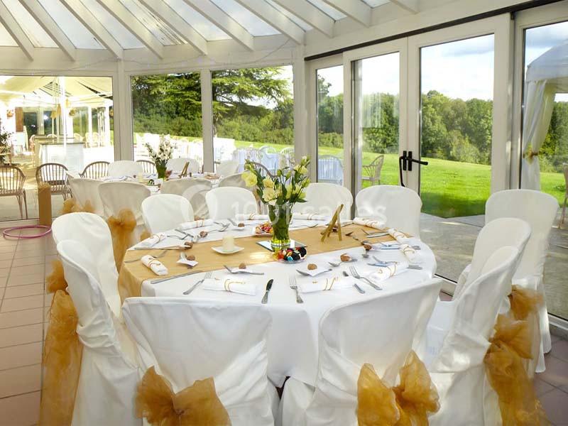 Salle lumineuse avec tables rondes dressées, nappes blanches, décorations dorées et vue sur un jardin verdoyant.