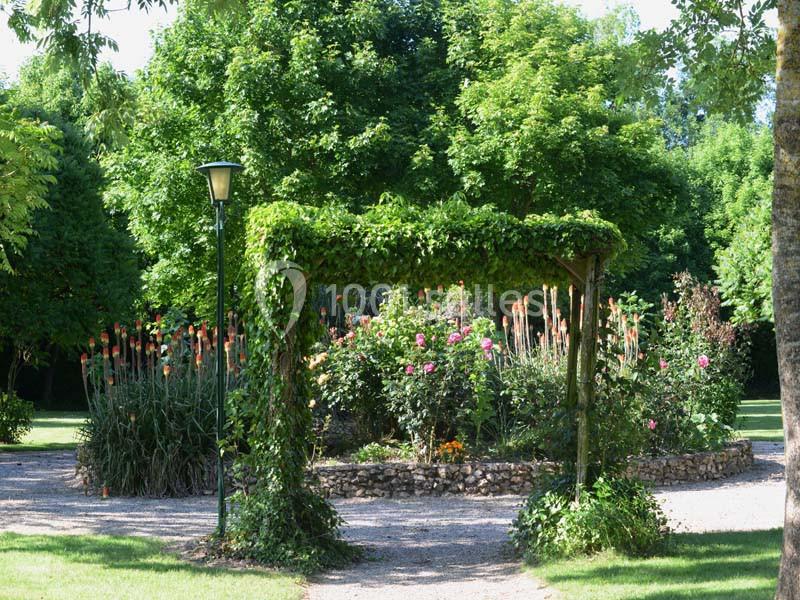 Pergola recouverte de verdure entourée de massifs fleuris dans un jardin arboré et ensoleillé.