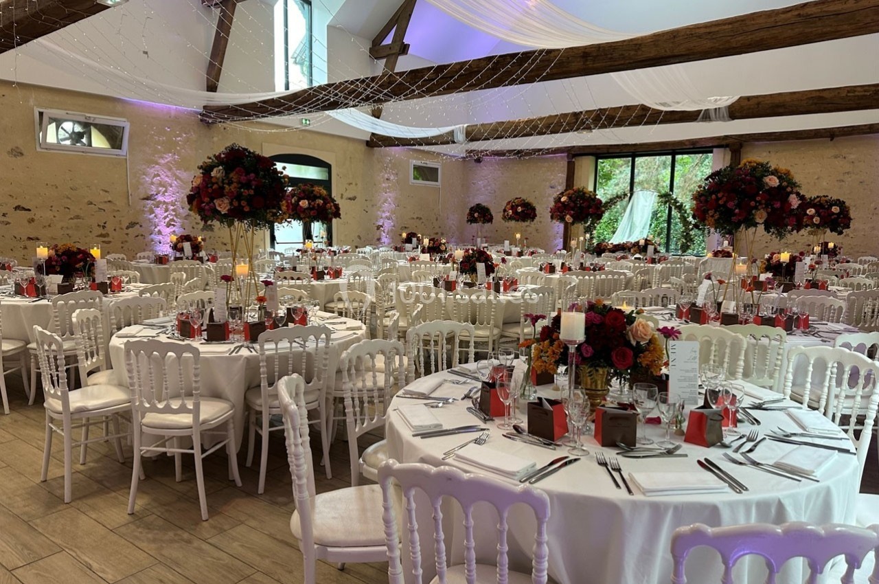 Salle de réception décorée pour un événement, avec tables rondes, chaises blanches et compositions florales colorées.