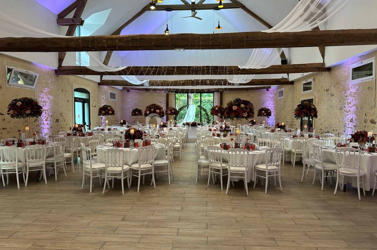 Salle de réception décorée avec des tables rondes, chaises blanches, fleurs rouges et éclairage tamisé.