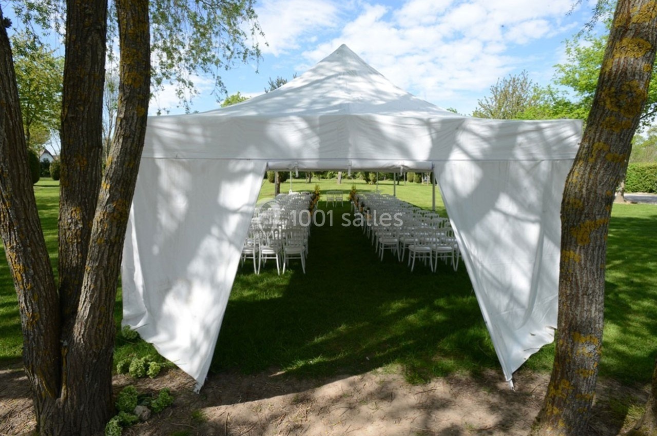 Tente blanche installée dans un jardin, abritant des rangées de chaises blanches alignées pour un événement en plein air.
