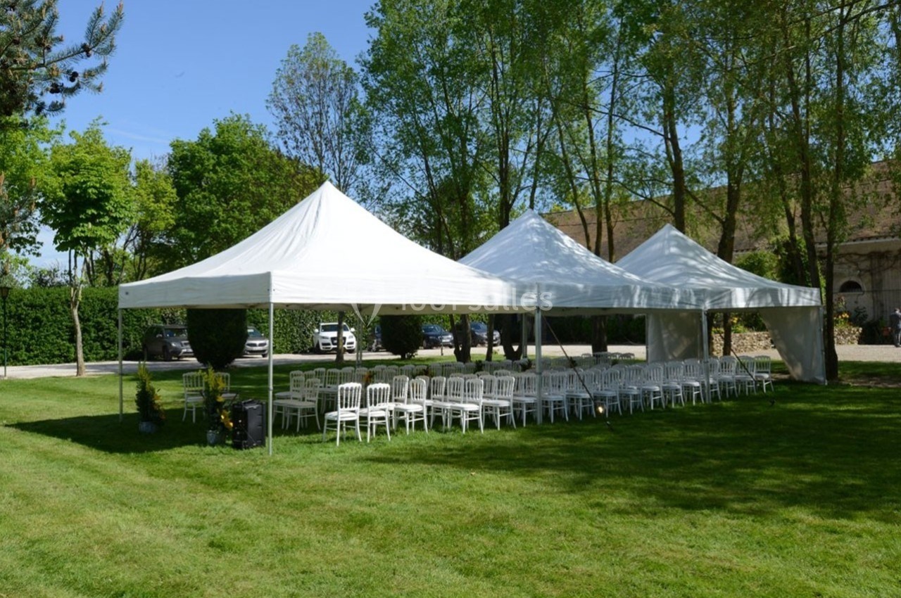 Tentes blanches montées sur une pelouse, entourées d'arbres, avec des rangées de chaises blanches disposées dessous.