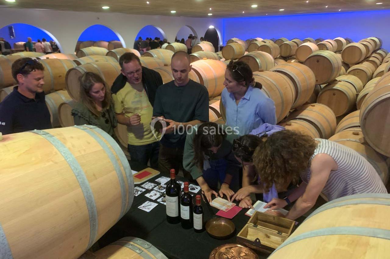 Location salle Moulis-en-Médoc (Gironde) - Château Maucaillou #7 Des personnes participent à une activité autour du vin dans une cave avec des tonneaux en bois en arrière-plan.