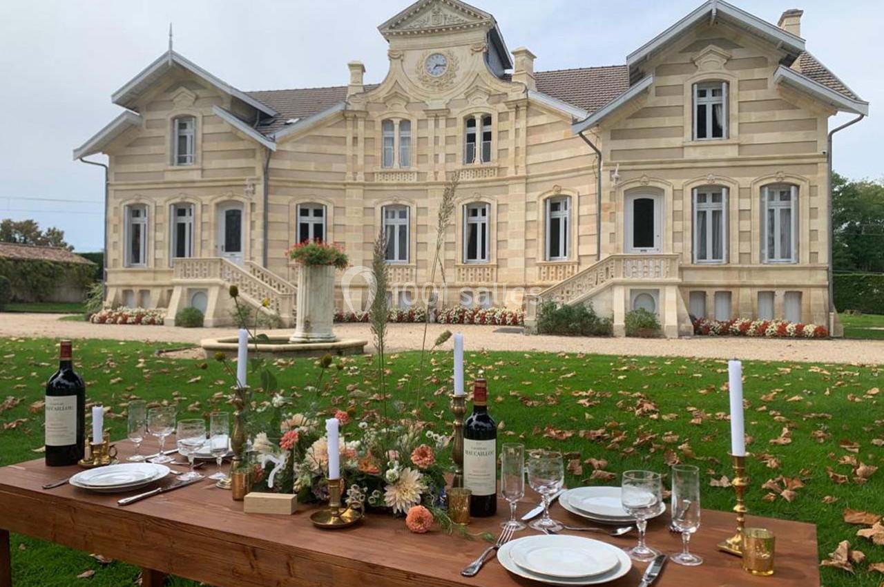 Location salle Moulis-en-Médoc (Gironde) - Château Maucaillou #22 Table dressée avec vaisselle et bouteilles de vin devant un manoir en pierre entouré de pelouse et de feuilles d'automne.