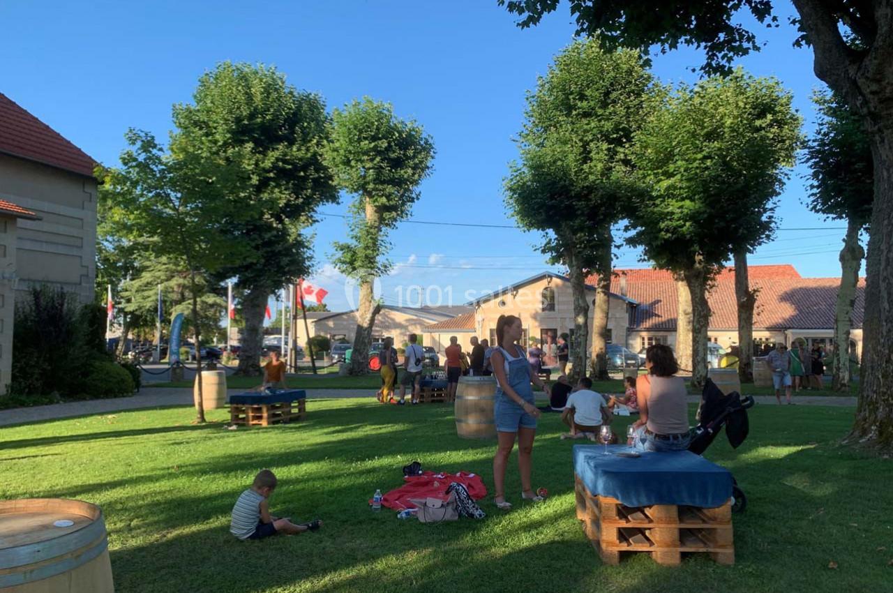 Location salle Moulis-en-Médoc (Gironde) - Château Maucaillou #16 Des personnes se détendent dans un parc ombragé avec des tables en palettes, entourées d'arbres et de bâtiments.