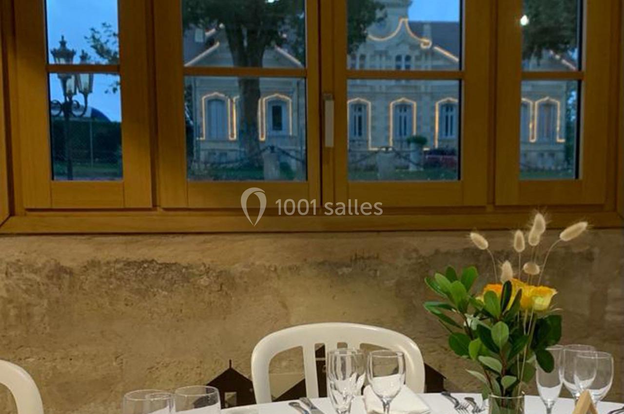 Location salle Moulis-en-Médoc (Gironde) - Château Maucaillou #21 Table dressée avec vaisselle et verres, décorée d'un bouquet, devant une fenêtre donnant sur un bâtiment éclairé.