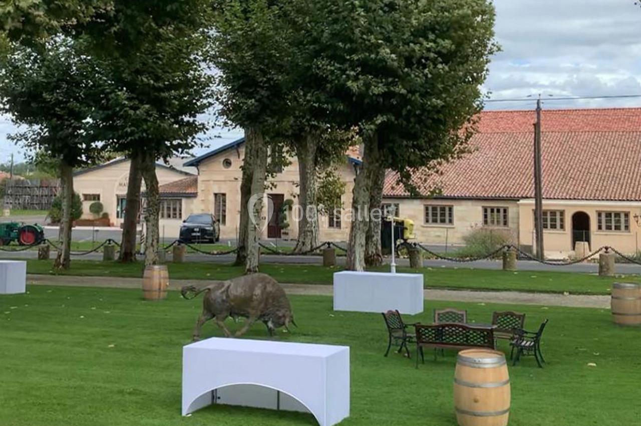 Location salle Moulis-en-Médoc (Gironde) - Château Maucaillou #15 Un jardin avec des tables, des tonneaux, une statue de sanglier et des bâtiments en arrière-plan.