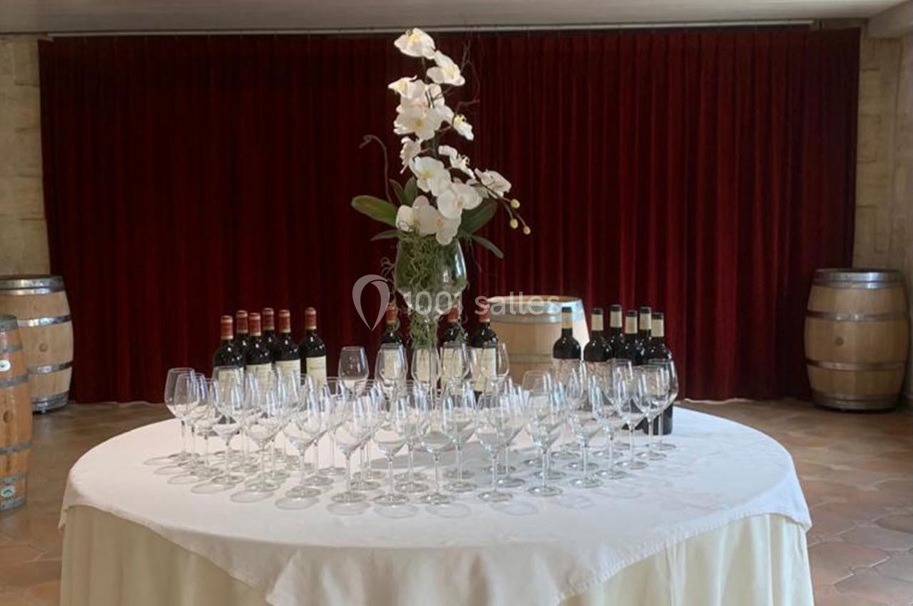Location salle Moulis-en-Médoc (Gironde) - Château Maucaillou #4 Table ronde avec nappes blanches, verres à vin alignés, bouteilles de vin et vase d'orchidées, devant un rideau rouge.