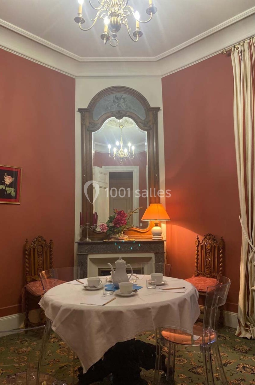 Salle à manger avec murs rouges, table ronde dressée, cheminée ornée d'un miroir et éclairage chaleureux.