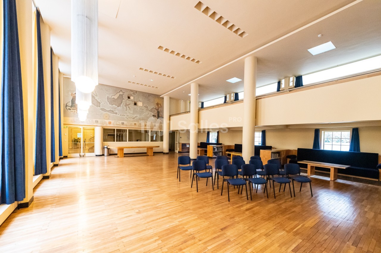 Salle spacieuse avec parquet en bois, chaises disposées, grandes fenêtres et fresque murale en arrière-plan.