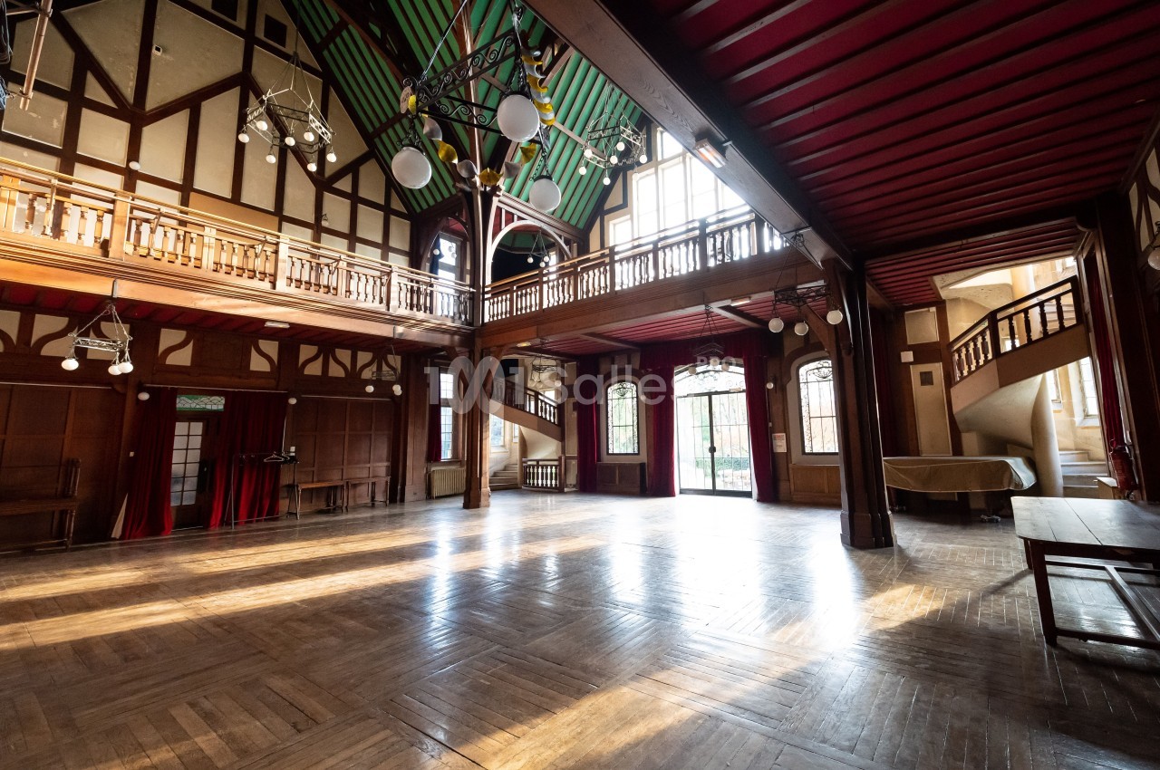 Grande salle lumineuse avec parquet en bois, balustrades, poutres apparentes et plafond coloré.
