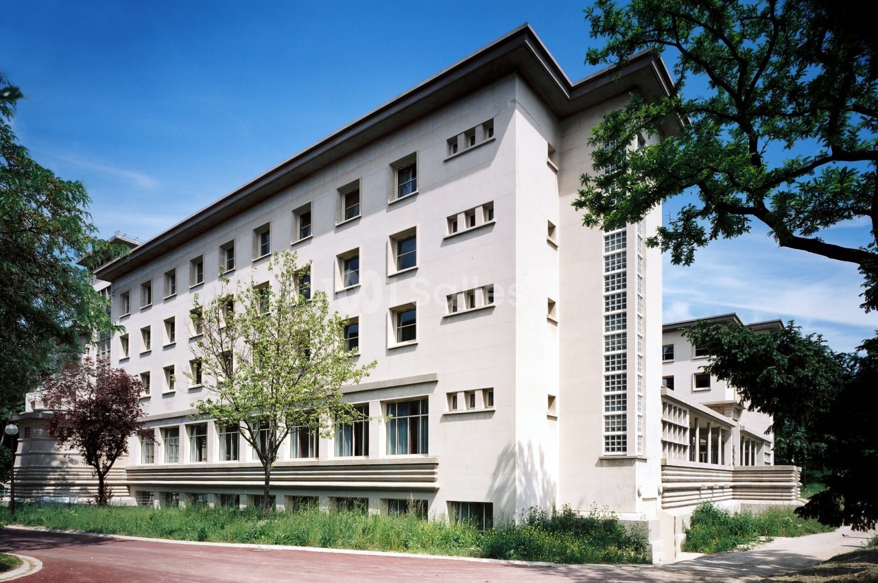 Bâtiment blanc à plusieurs étages avec des fenêtres rectangulaires, entouré d'arbres et de végétation.