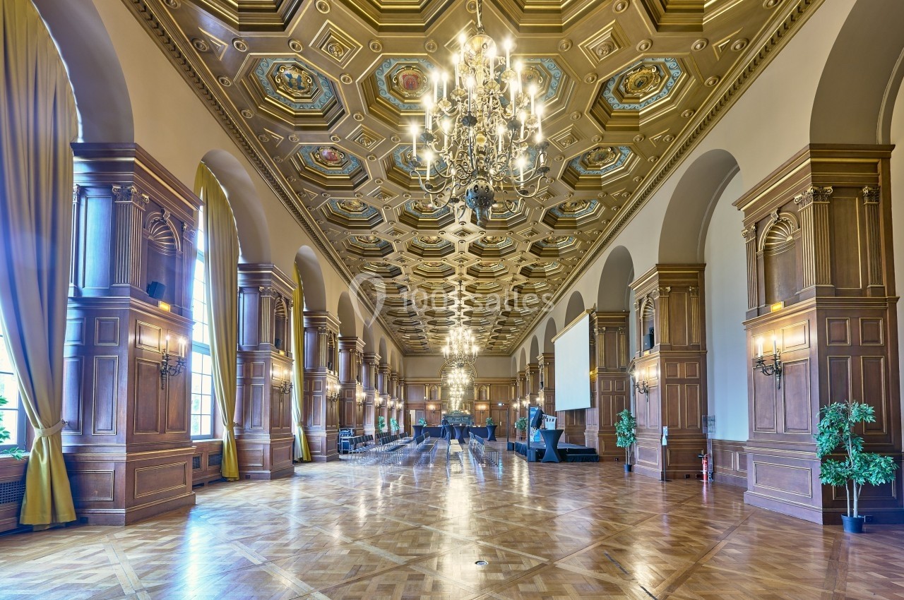 Grande salle avec parquet, hauts plafonds ornés, lustres imposants, colonnes en bois et rideaux encadrant des fenêtres.