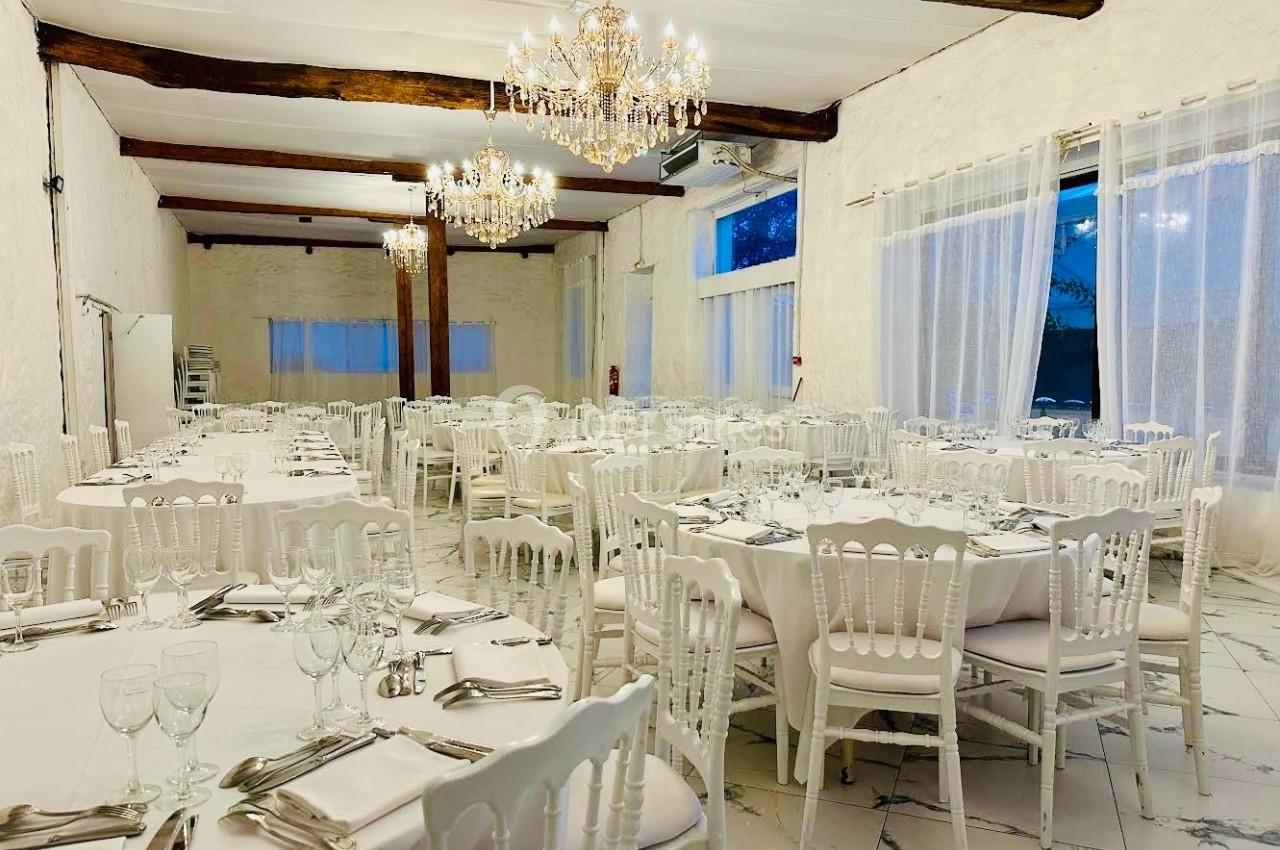 Salle de réception lumineuse avec tables rondes dressées, chaises blanches et lustres suspendus.