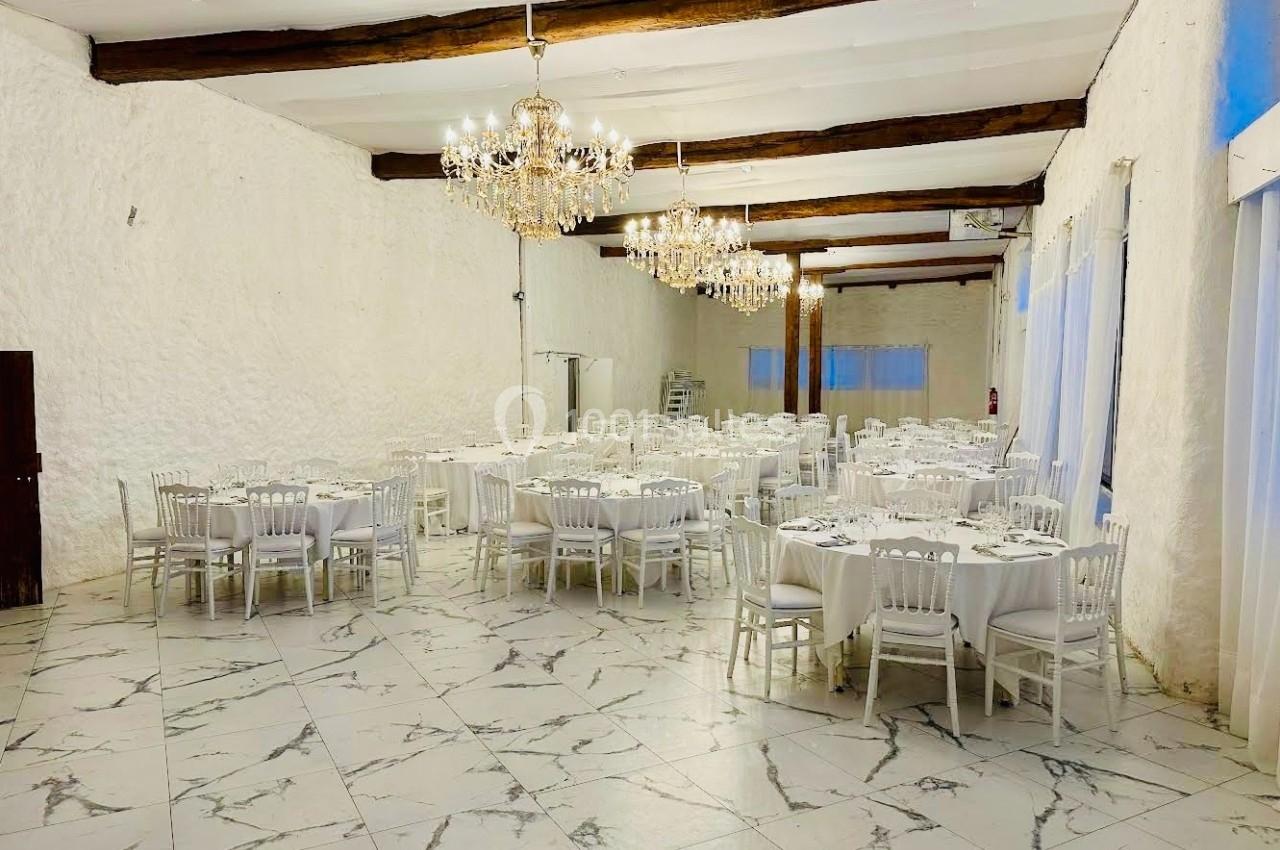 Salle de réception lumineuse avec tables rondes dressées, chaises blanches et lustres suspendus au plafond.