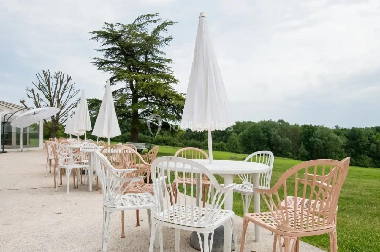 Terrasse avec tables et chaises blanches et roses, parasols ouverts, donnant sur un espace vert arboré.