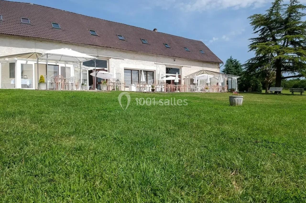 Bâtiment en pierre avec toit en pente, terrasse aménagée avec parasols, entouré d'une pelouse et d'arbres.
