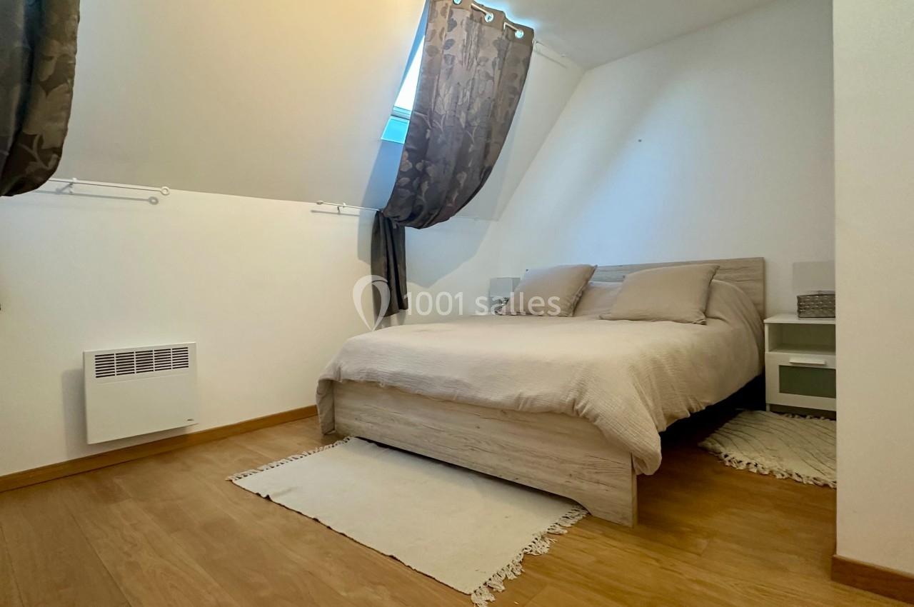 Chambre lumineuse avec lit double, parquet, murs blancs, rideaux sombres et petite fenêtre inclinée.