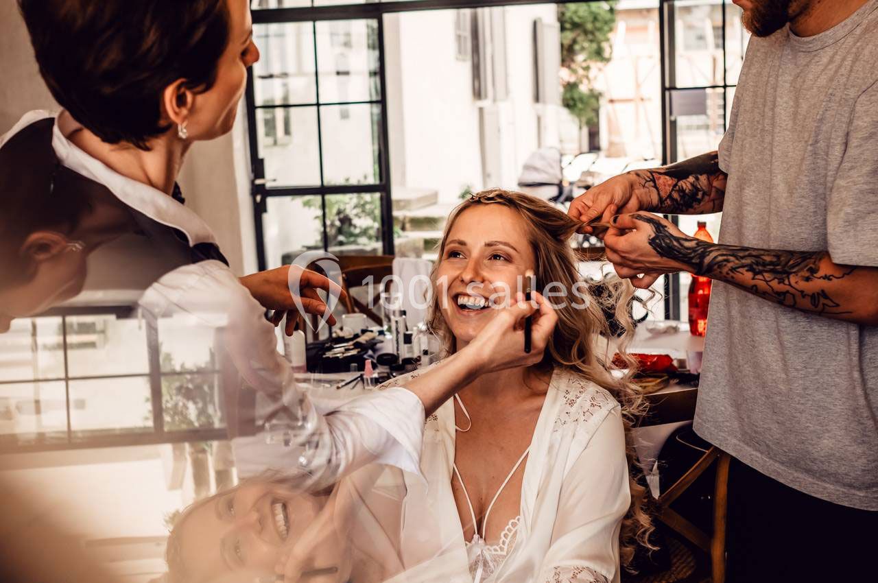 Une femme souriante se fait coiffer et maquiller par deux professionnels dans une pièce lumineuse.