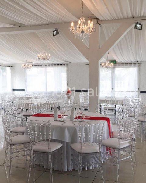 Salle de réception lumineuse avec tables rondes dressées, chaises transparentes et lustres suspendus.