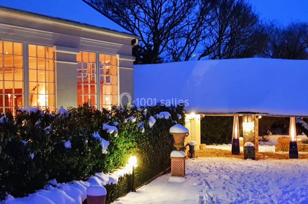 Bâtiment éclairé avec de grandes fenêtres, entouré de neige et de végétation, sous un ciel bleu en soirée.