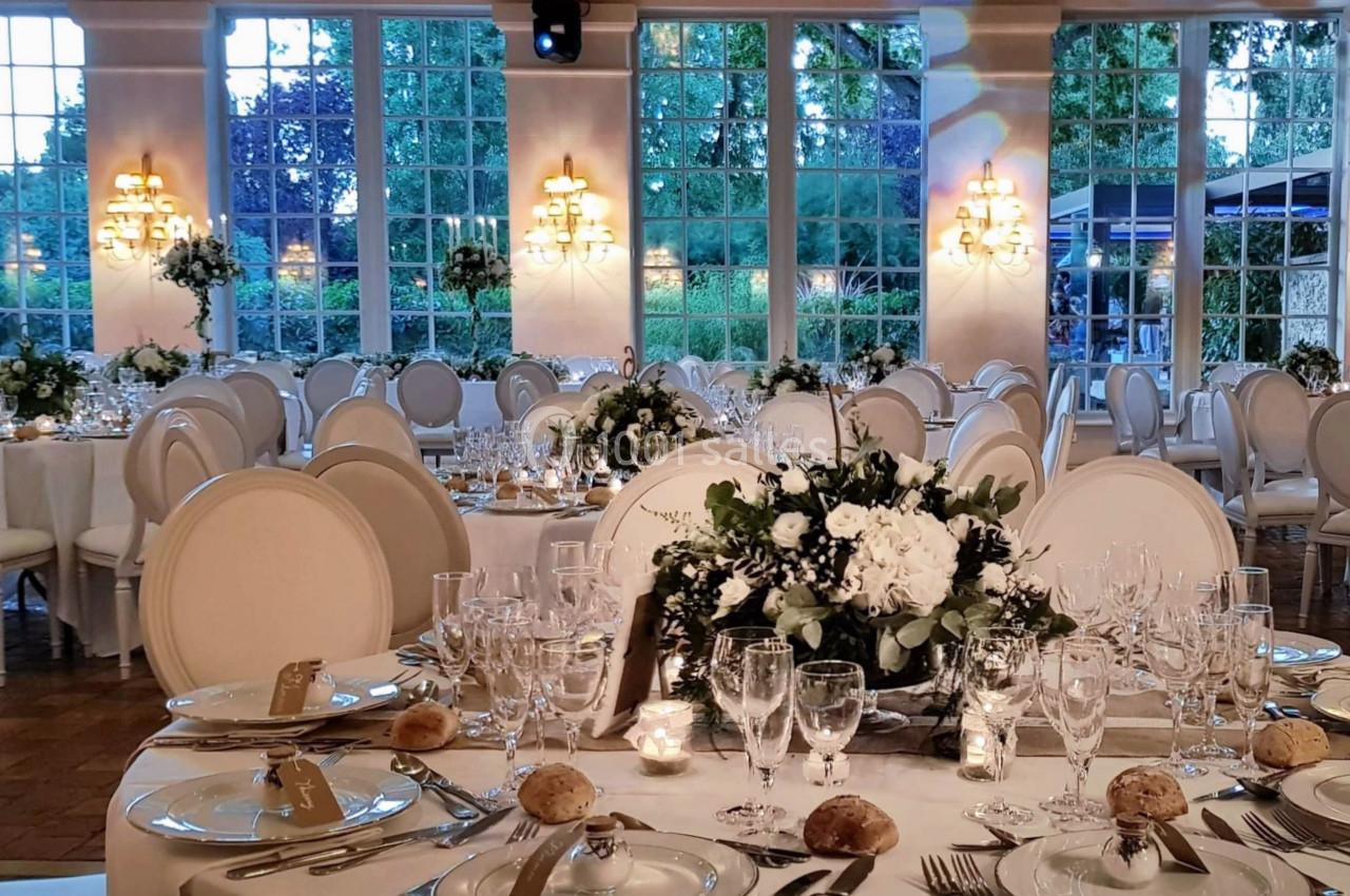 Salle de réception élégante avec tables rondes dressées, décorées de fleurs blanches et éclairées par des lustres.