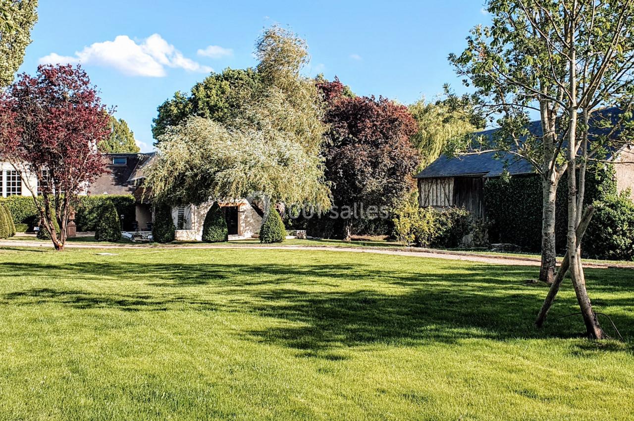 Grand jardin verdoyant avec pelouse, arbres variés et bâtiments en pierre entourés de haies.