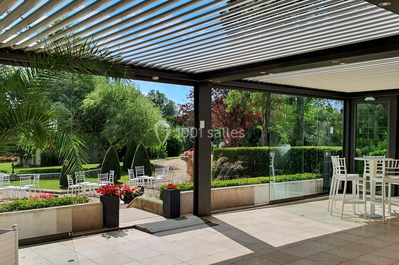 Terrasse couverte avec pergola à lames orientables, vue sur un jardin arboré et mobilier de jardin blanc.