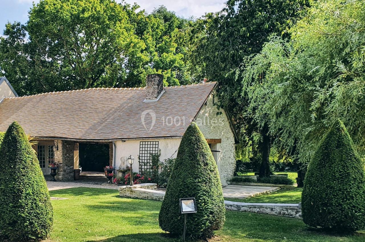 Maison en pierre avec toit en tuiles, entourée d'arbres, de buissons taillés et d'un jardin verdoyant.