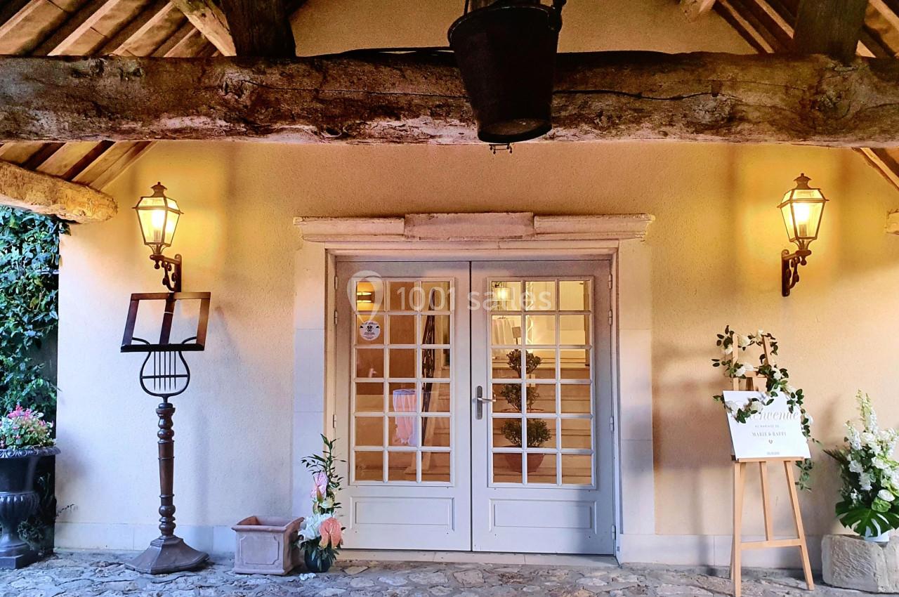 Entrée d'un bâtiment avec porte vitrée, encadrée de deux lanternes murales et décorée de plantes et d'un chevalet.