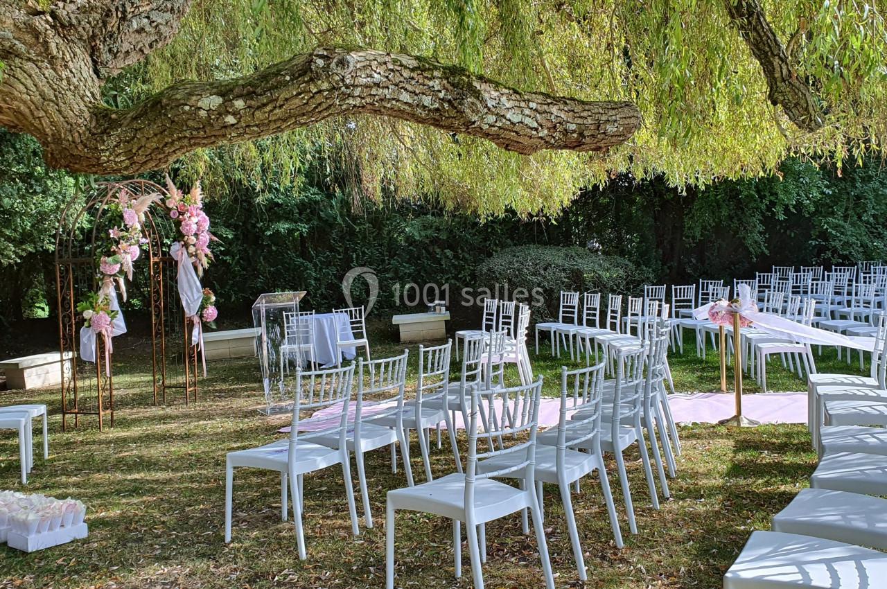 Chaises blanches disposées en extérieur sous un arbre, avec une arche fleurie et une allée décorée pour une cérémonie.