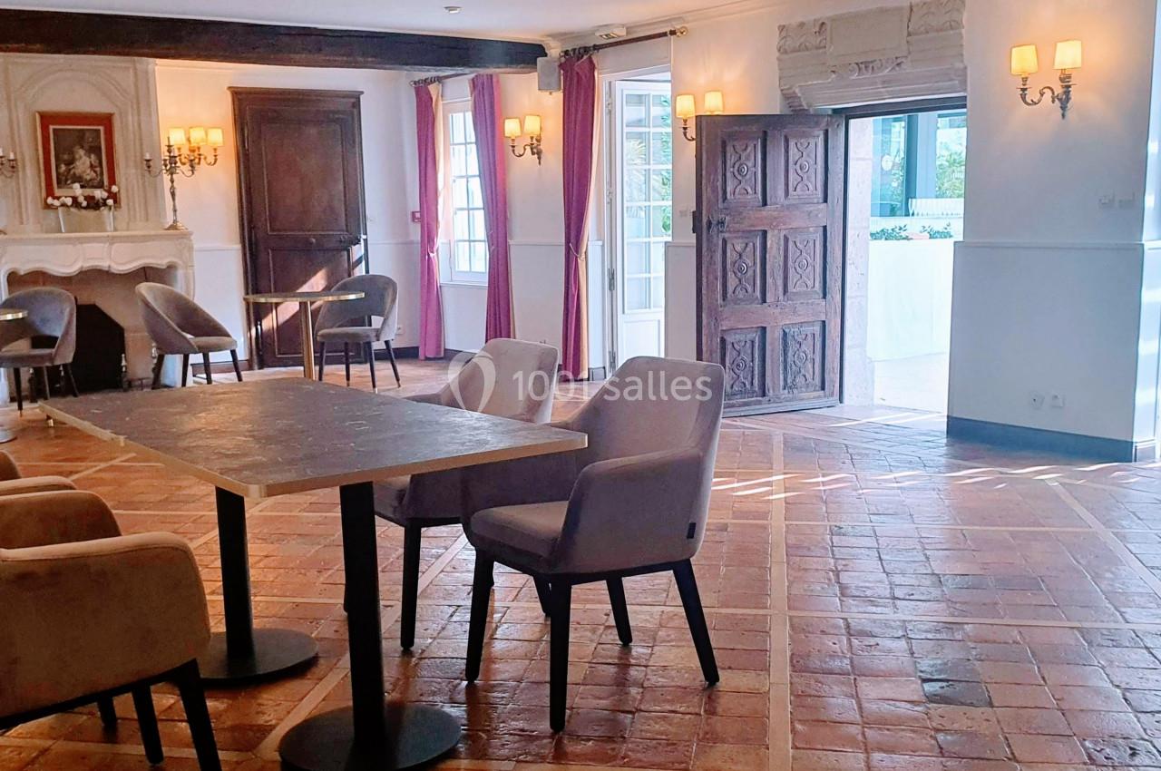 Salle lumineuse avec carrelage en terre cuite, tables et chaises, murs blancs, cheminée et grandes ouvertures sur l…