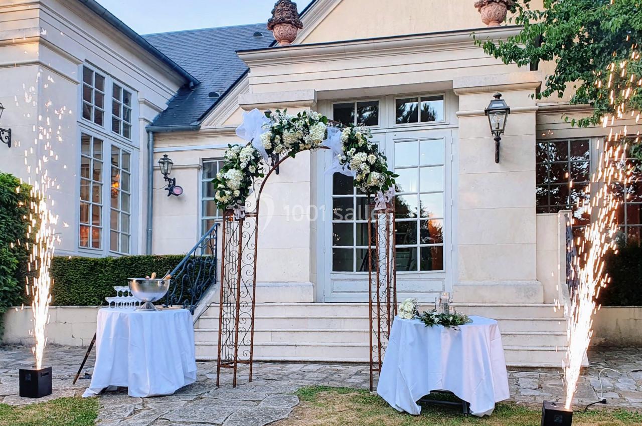 Arche décorée de fleurs blanches devant une maison élégante, entourée de tables et de jets d'étincelles.