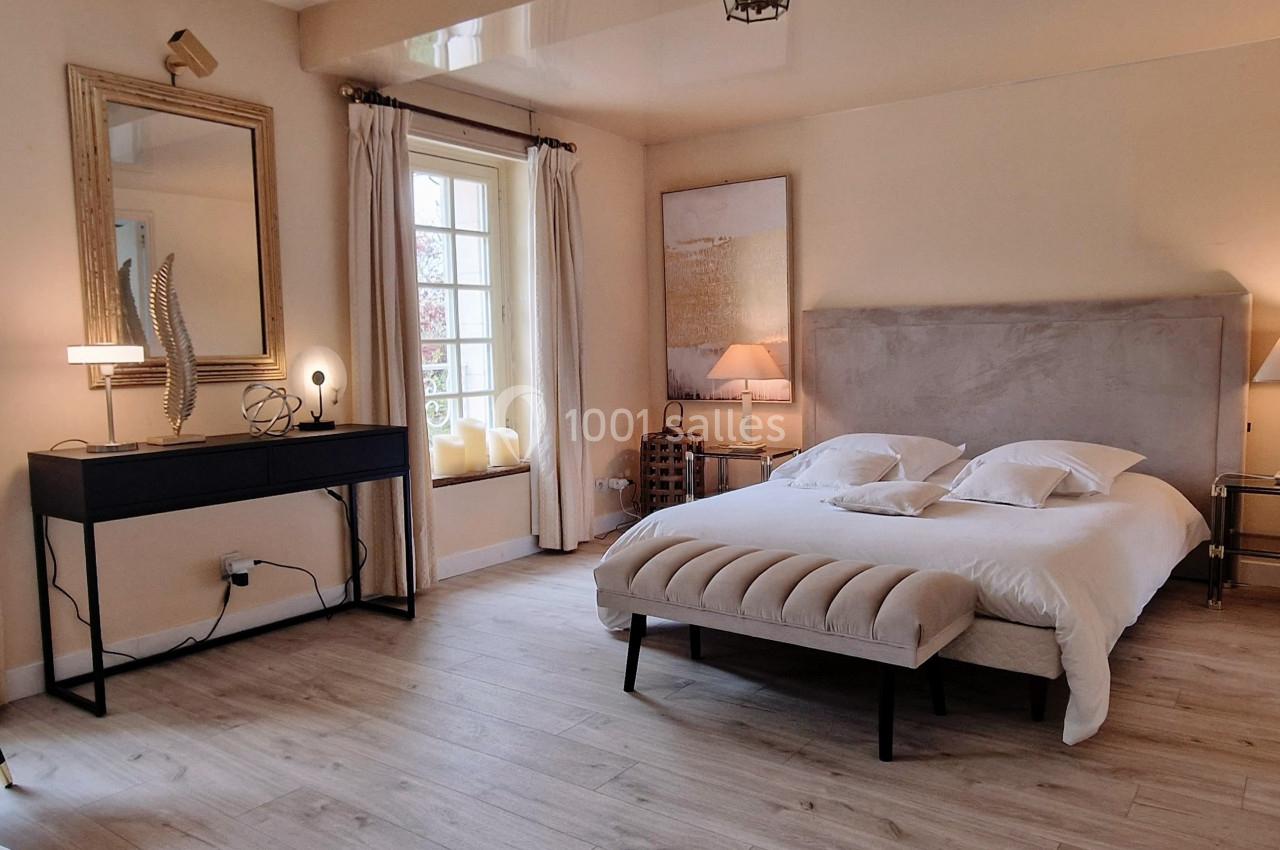 Chambre lumineuse avec lit double, bureau noir, fauteuils, miroirs et grandes fenêtres donnant sur l'extérieur.