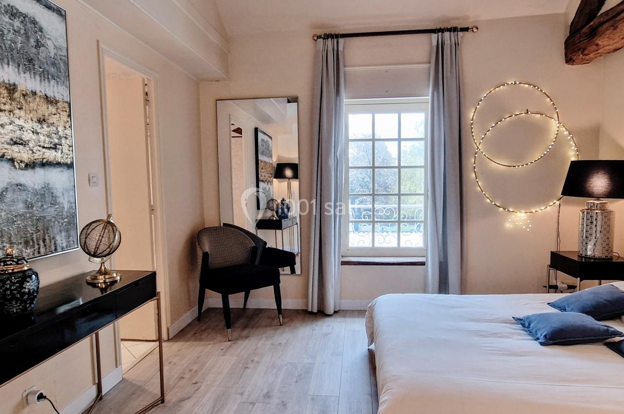 Chambre lumineuse avec lit double, miroir, bureau, fauteuil, et décoration murale, vue sur une fenêtre avec rideaux.