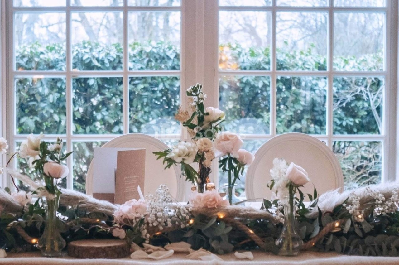 Table décorée avec des fleurs pastel, feuillages et bougies, placée devant une grande fenêtre donnant sur un jardin.