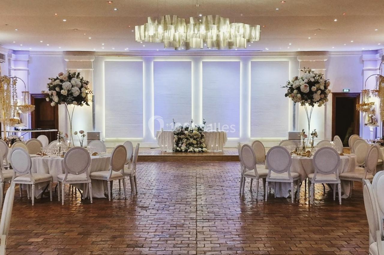 Salle de réception élégante avec tables rondes dressées, chaises blanches et décorations florales raffinées.