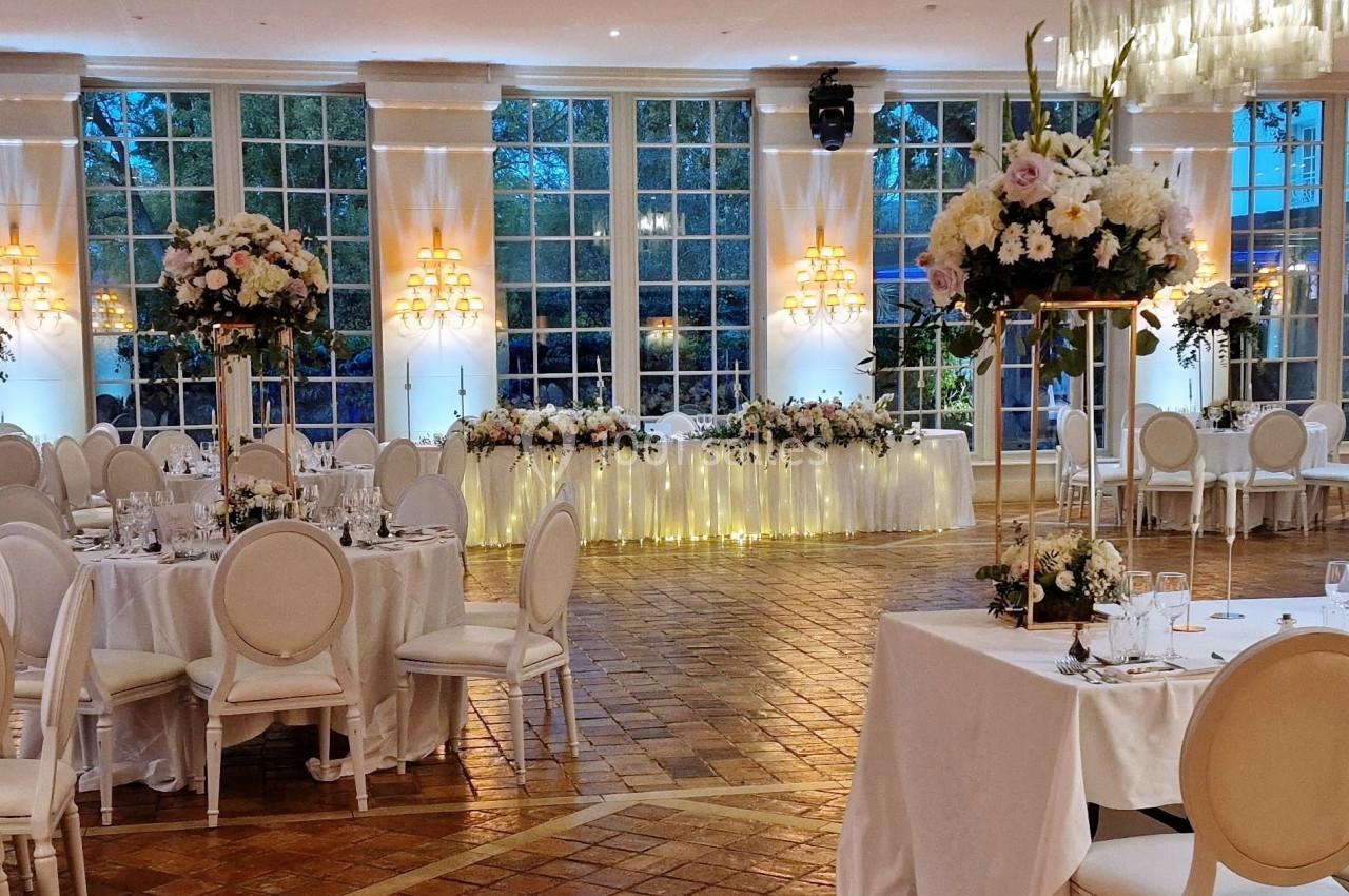 Salle de réception élégante avec tables dressées, chaises blanches et grandes compositions florales, éclairée par des baies…