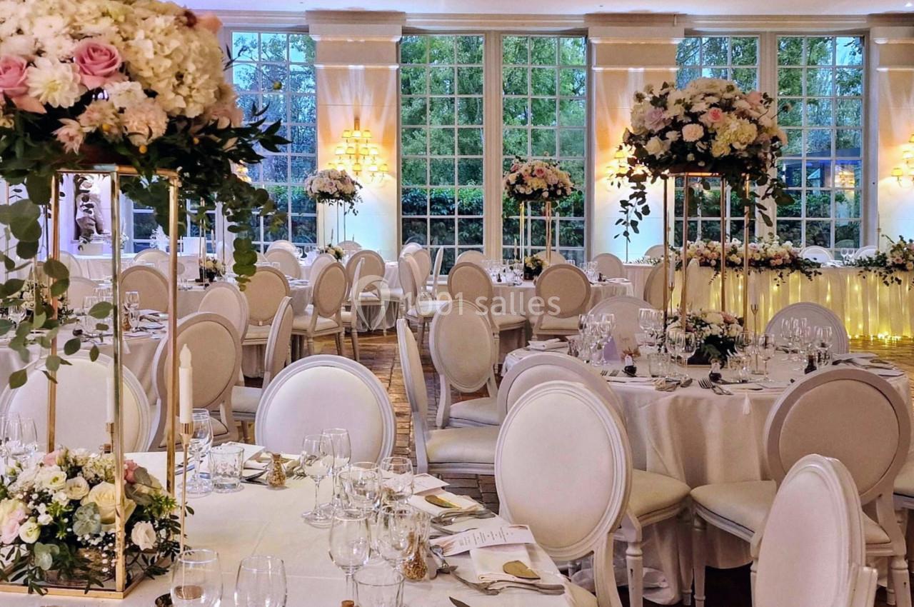 Salle de réception élégante avec tables rondes décorées de fleurs blanches et roses, éclairée par de grandes fenêtres.