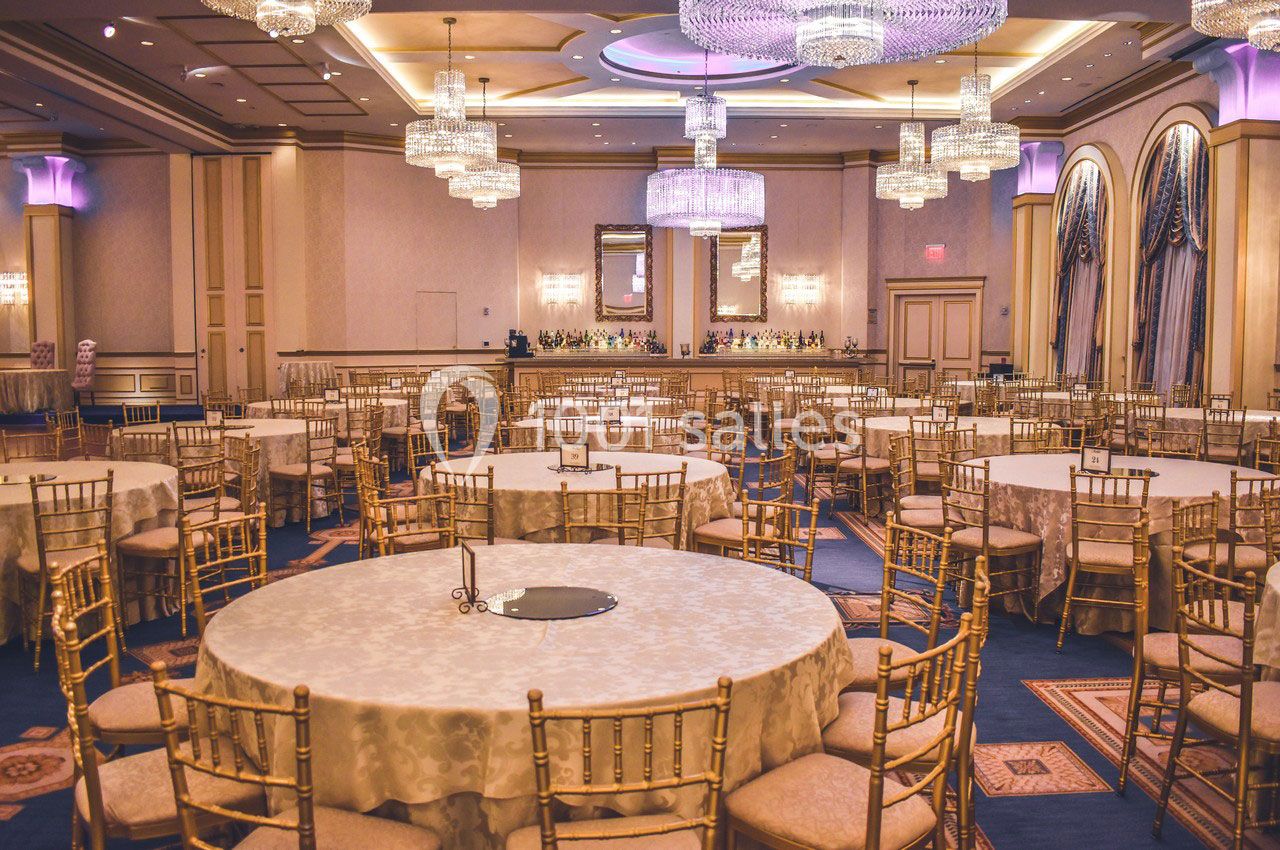 Salle de réception élégante avec tables rondes dressées, chaises dorées et lustres imposants.