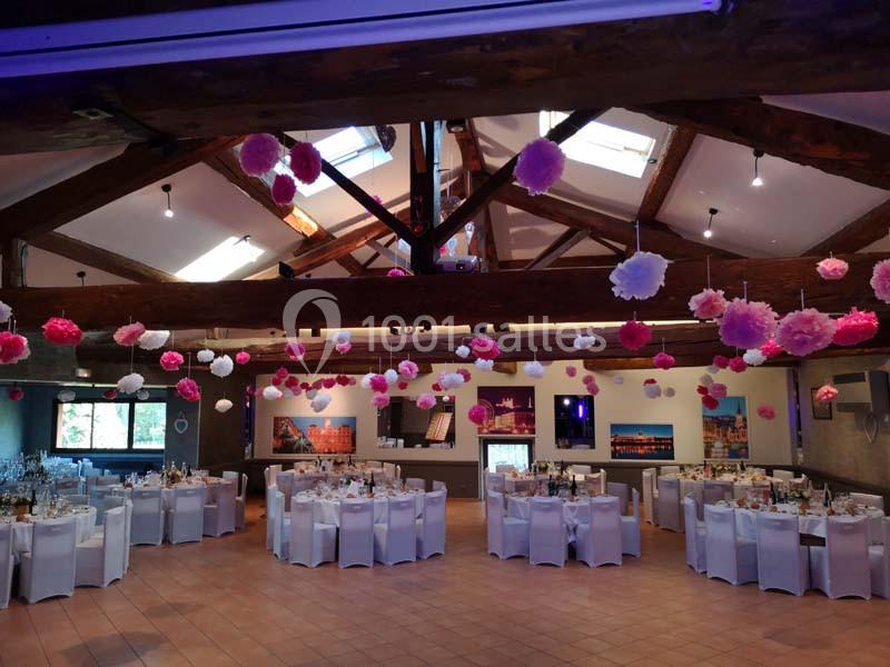 Salle de réception décorée avec des tables dressées et des suspensions colorées sous une charpente en bois apparente.