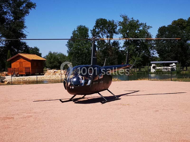 Hélicoptère noir posé sur un terrain en gravier, avec un étang et une cabane en bois en arrière-plan.